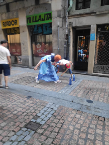 A woman and man with colourful stockings over their head, trying to knock over bottles of water with the stockings.