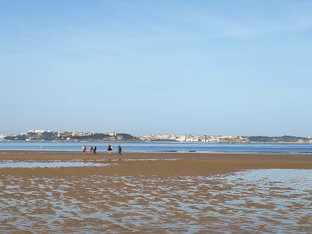 A group of pilgrims walking along the beach, photographed from the side, framed by the sea.