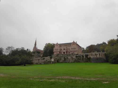 A manor house with reddish stone. A church next door seems to be made from similar stone. Grey clouds, and a grassy lawn in the foreground.