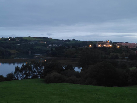 Dawn scene of San Vicente de la Barquera. Stone buildings, orange lights, and a dark lake in between.