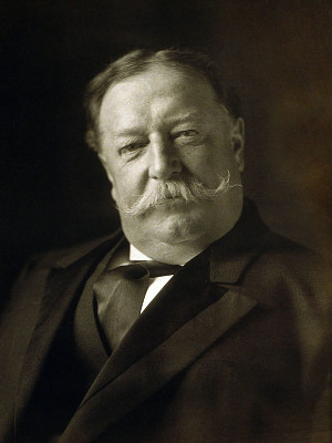 A picture of President Taft, in black & white. He appears to be a good-humoured man. He has a moustache. He's sitting in a chair. He's rotund.